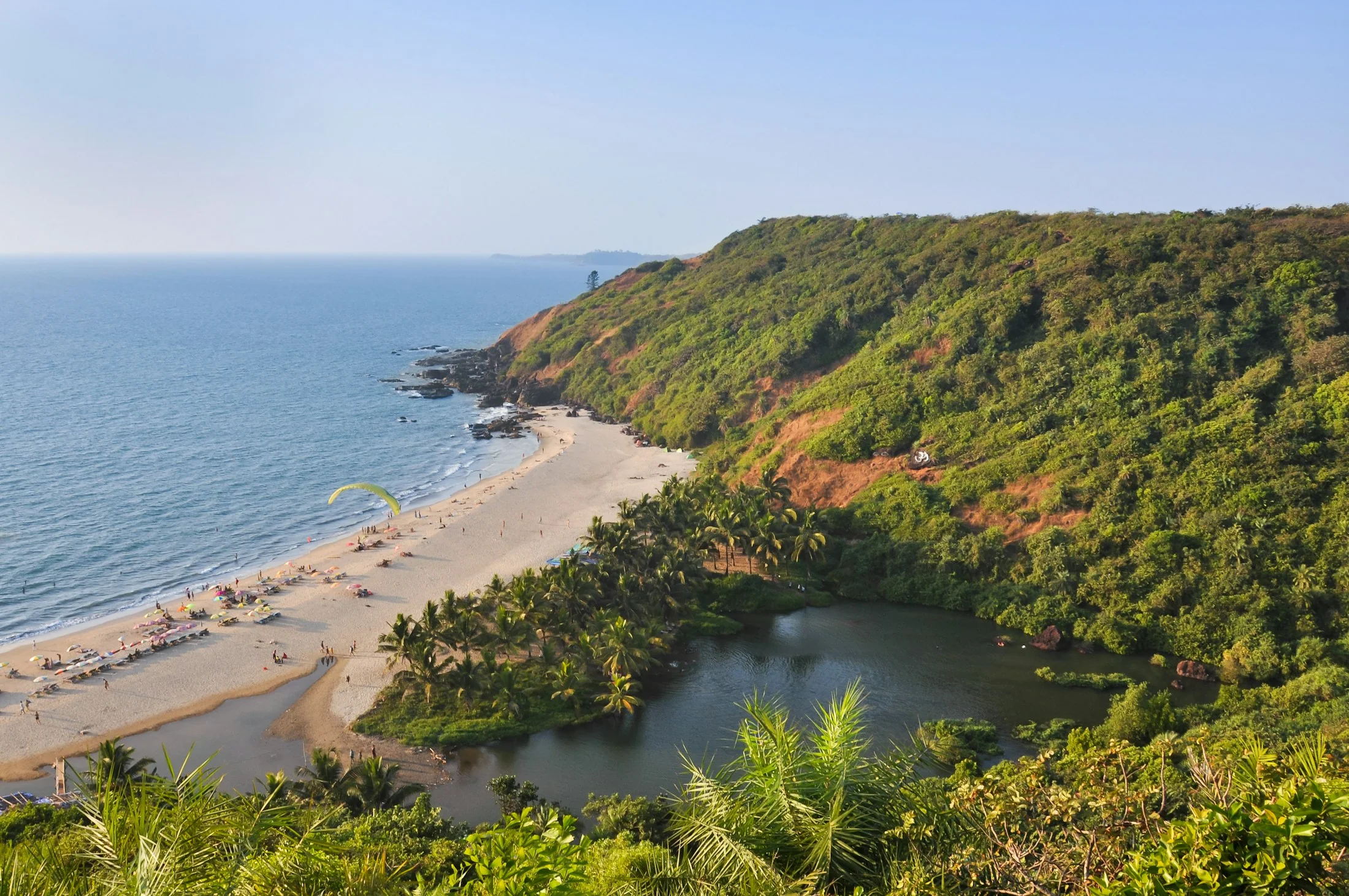 A curved sandy cove on the Goan coast seen from a forested headland above, surf running in along the beach